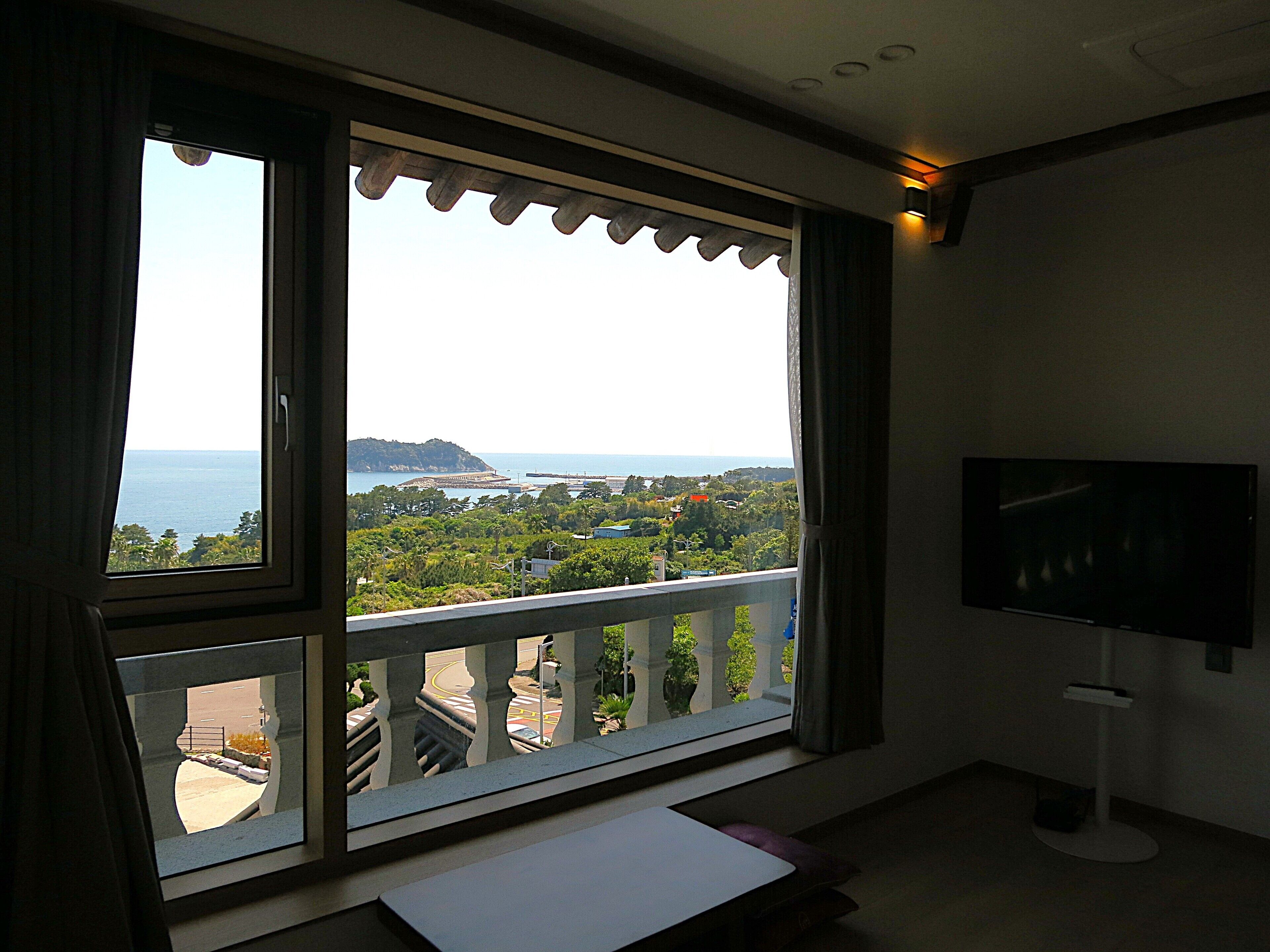 A room with a large window offering a view of the sea and a distant island. There is a TV mounted on the wall.