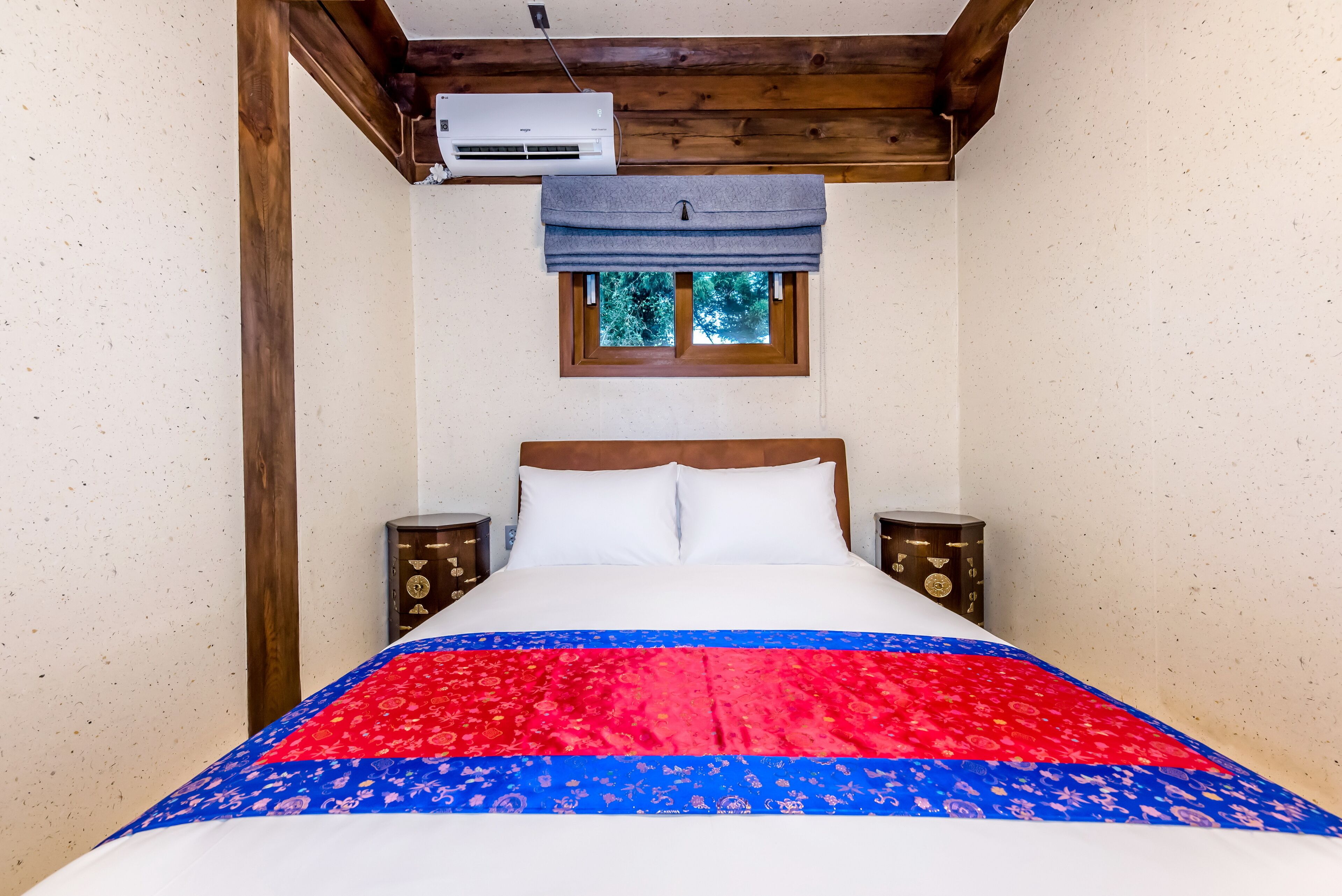 Un dormitorio con cama, cabecero de madera, dos mesitas de noche, una ventana con persiana azul y un equipo de aire acondicionado en la pared.