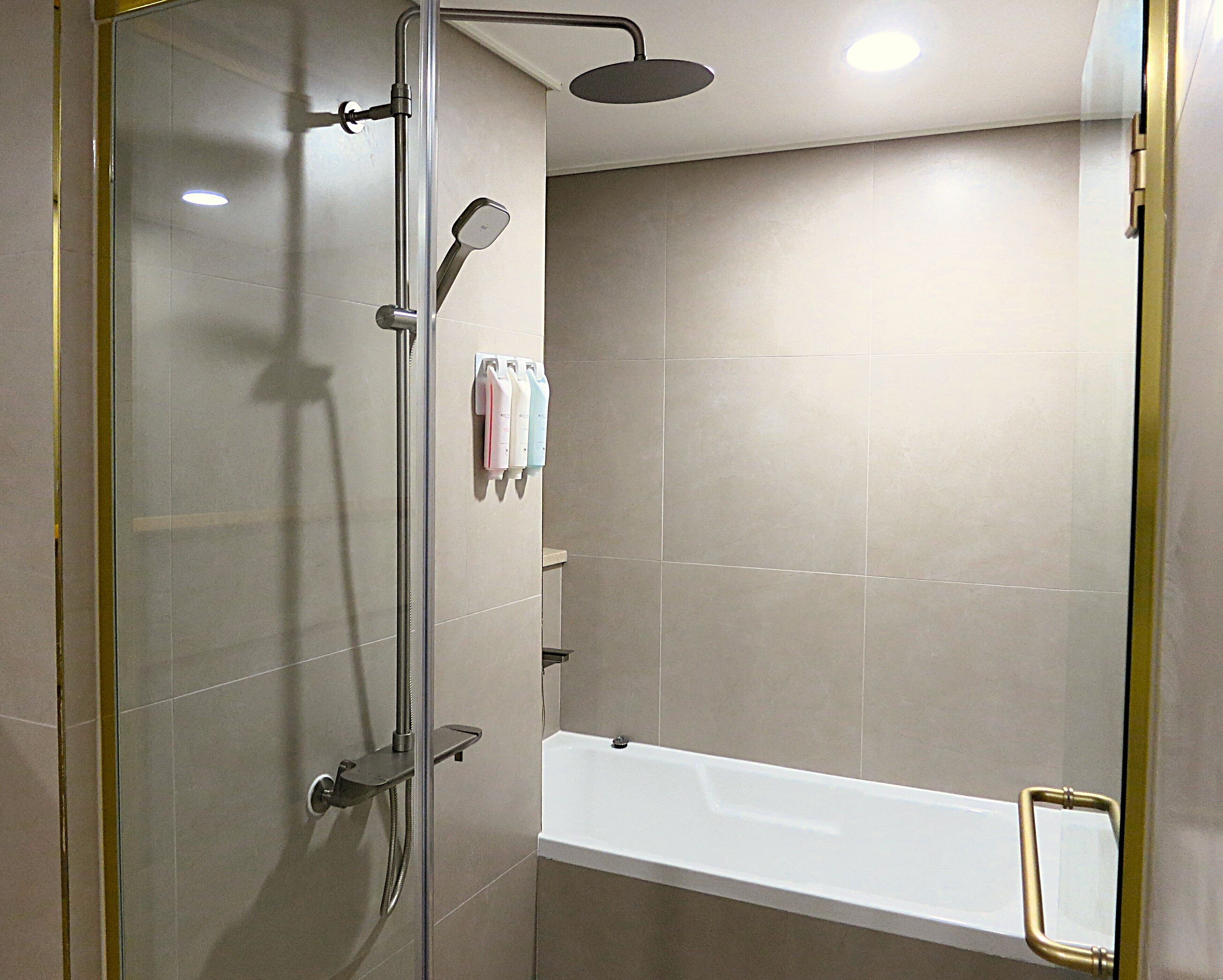 A modern shower area with a glass door, a handheld showerhead, a wall-mounted faucet, and a white bathtub with a gold grab bar.