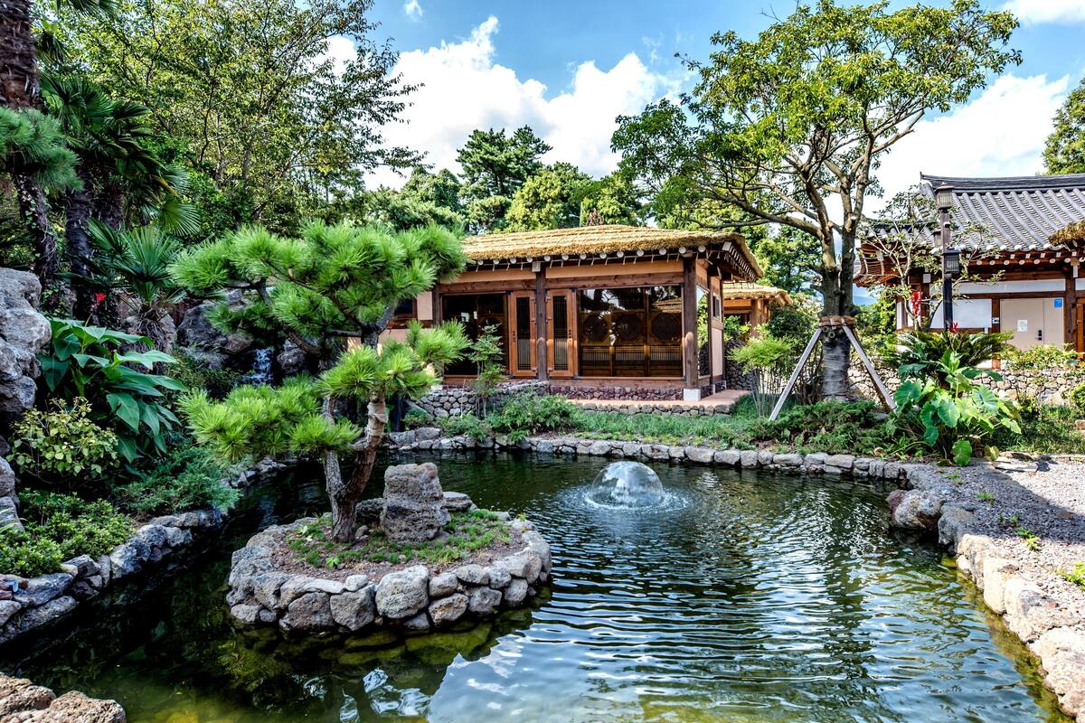 Un edificio tradicional japonés con estructura de madera y tejado de tejas, rodeado de abundante vegetación y un pequeño estanque con un puente de piedra.