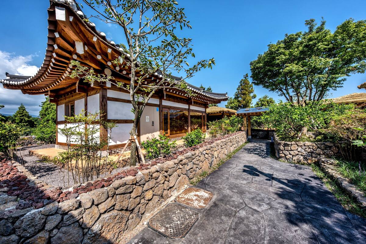 Edificio japonés tradicional con techo curvo y vigas de madera, rodeado de un sendero de piedra y abundante vegetación.