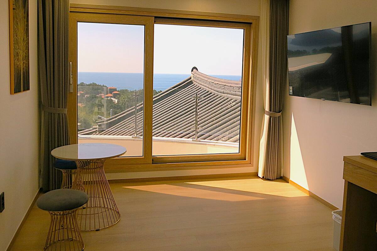 Una habitación de hotel moderna con un amplio ventanal corredero que da a un balcón, una mesa redonda con dos sillas, un televisor de pantalla plana montado en la pared y suelo de madera.