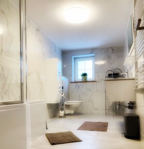 A bathroom with a shower, toilet, and sink, marble walls, and a window.