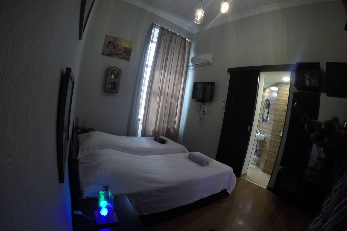A hotel room with a bed, a bedside table with a blue light, a TV mounted on the wall, and a door leading to a bathroom.