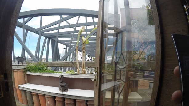 A view through a circular window of a terrace with terracotta planters, a wooden bench, and a metal sculpture. - Hotel Imaginarium (Tbilisi)