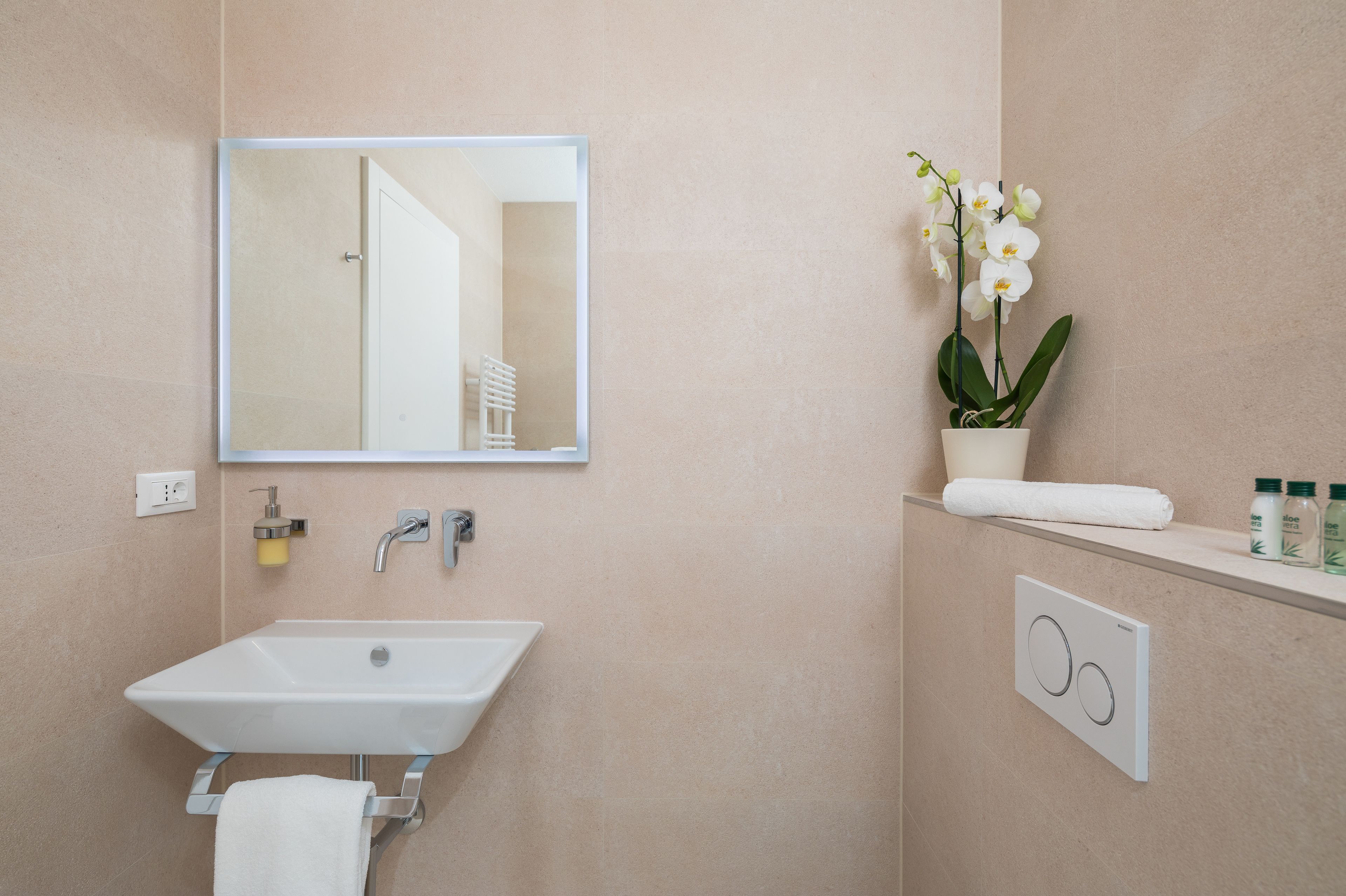 Un bagno con un lavandino bianco, uno specchio, un portasciuga e un'orchidea in vaso, con un asciugamano sul ripiano.