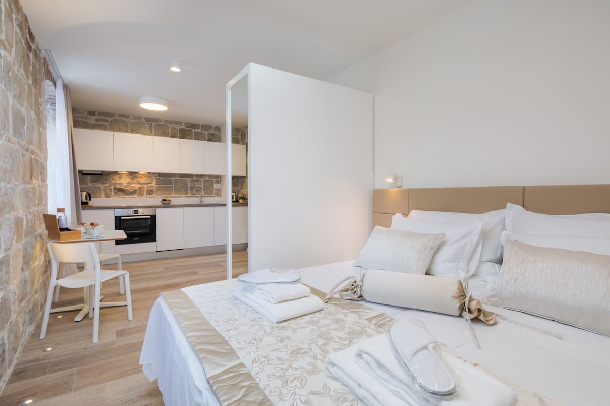 A modern hotel room with a neatly made bed, a small dining table, and a kitchen area in the background.