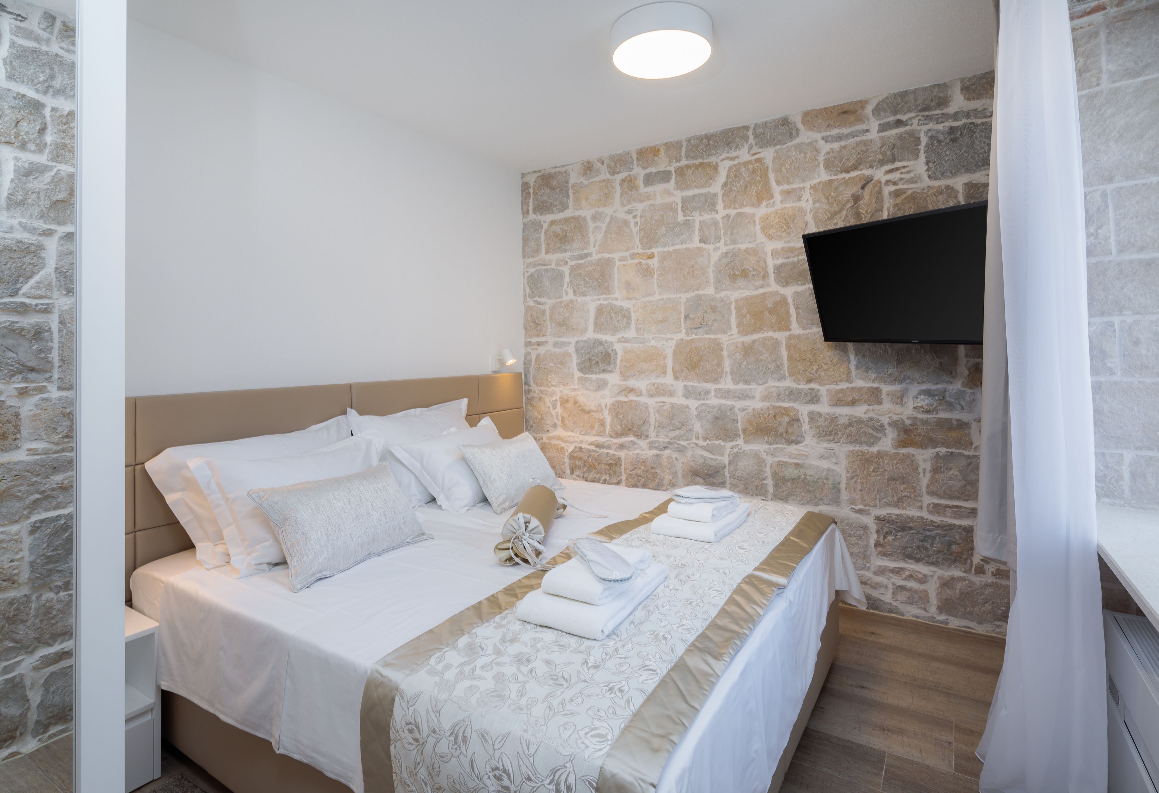 A bedroom with a stone wall, a flat-screen TV mounted above the bed, and neatly folded towels on the bed.