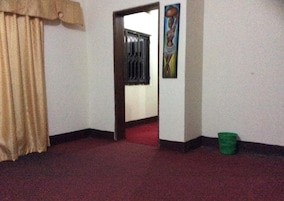 Una habitación con alfombra roja, cortina color beige, un bote de basura verde y una puerta que da a otra habitación.