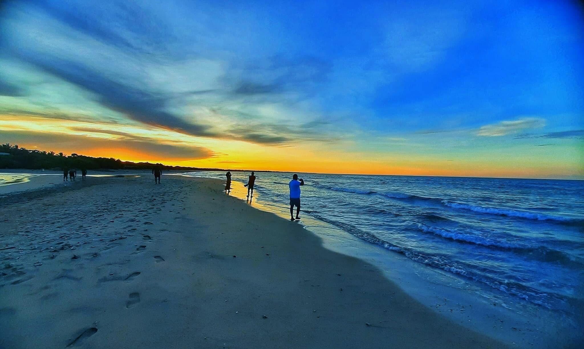 En strand med fodspor, der fører hen til vandet, en klar himmel og en solnedgang.