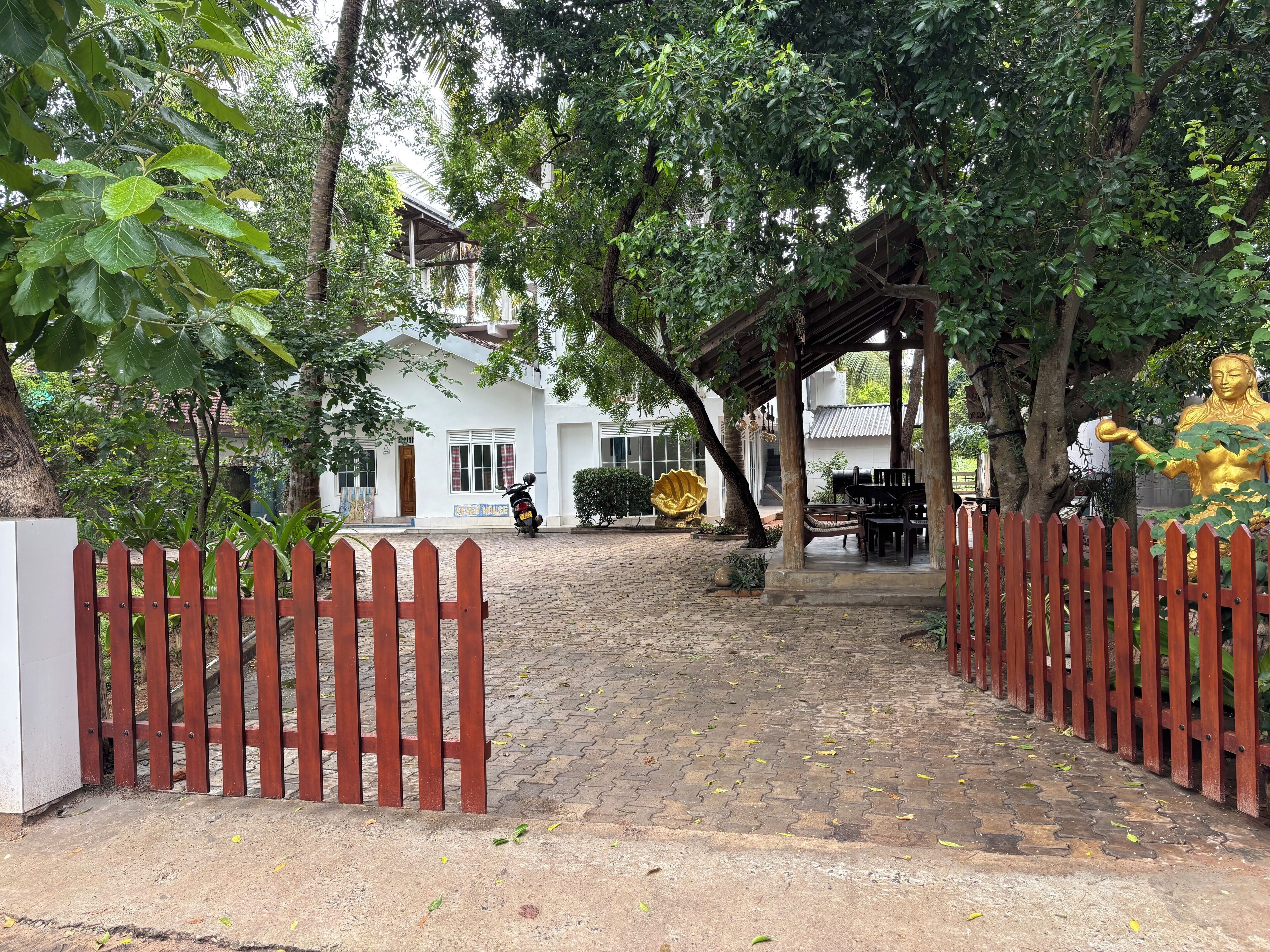 Una entrada protegida por una reja, con un camino empedrado que lleva a un edificio con una zona de estar cubierta y una estatua dorada.