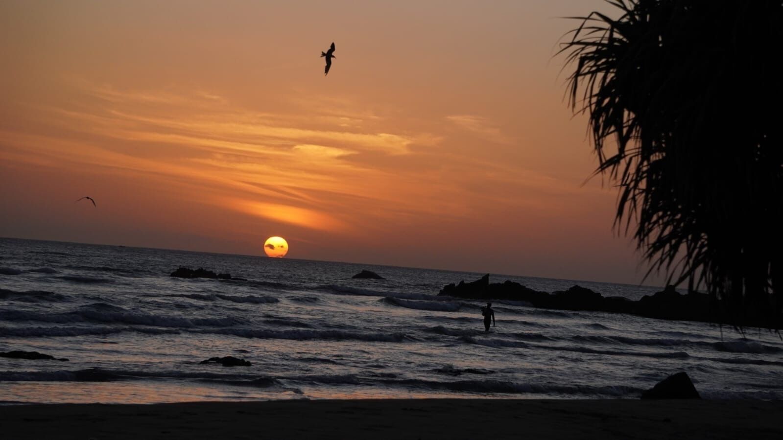 Un atardecer sobre el océano con olas y la silueta de una persona caminando.