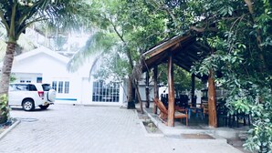 A covered outdoor seating area with tables and chairs, a white building in the background, and a parked white SUV.