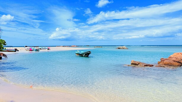 A beach with clear turquoise water, a boat, and rocky outcrops.