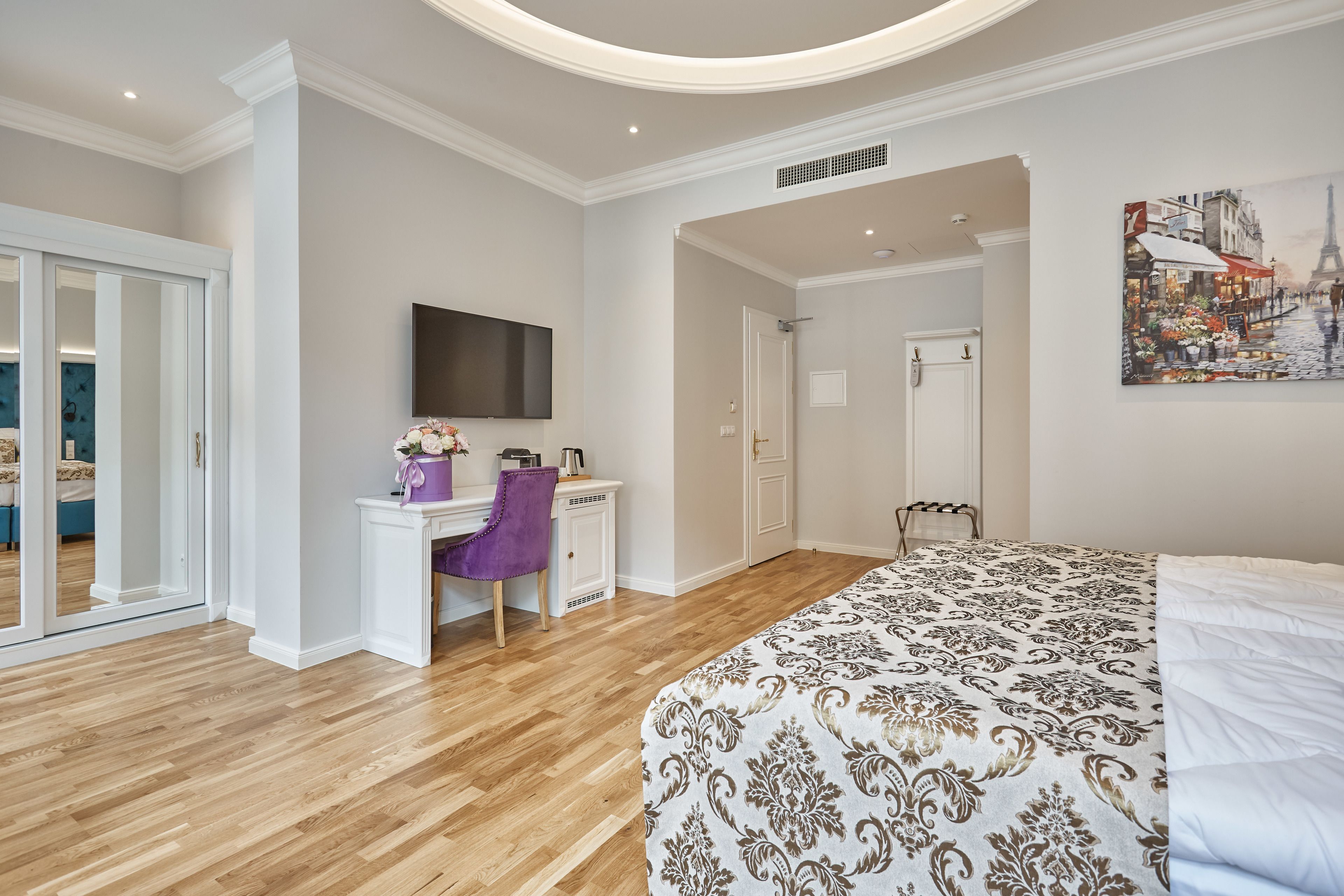 A modern bedroom with a large bed, a desk with a chair, a flat-screen TV, and a door leading to another room.