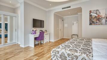 A modern bedroom with a large bed, a desk with a chair, a flat-screen TV, and a door leading to another room.