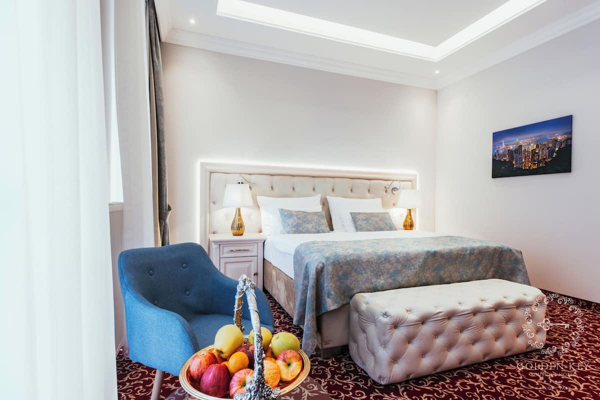 Habitación de hotel con cama, un sillón azul, una cesta de fruta y un cuadro con una vista urbana.