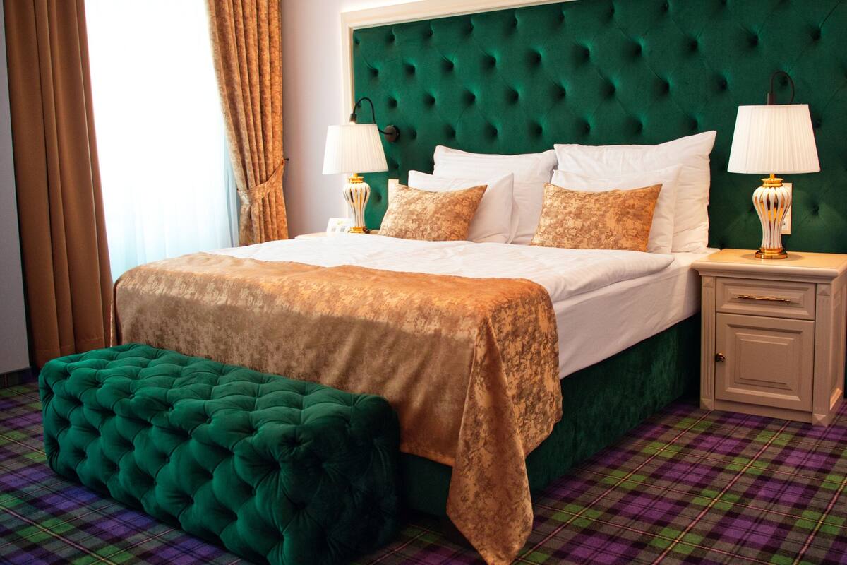 Habitación de hotel con cabecera tapizada verde con diseño capitonado, cama grande con ropa de cama blanca, dos lámparas de noche, un banco tapizado en verde y suelo con estampado de cuadros.