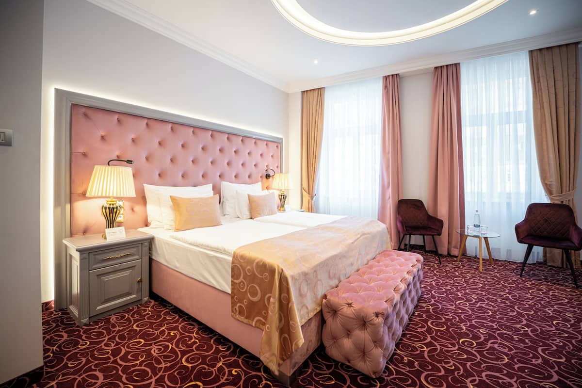 Habitación de hotel con una cama grande, una mesita de noche con lámpara, un banco, dos sillas y una pequeña mesa.