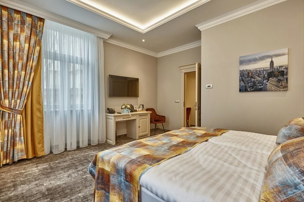 A hotel room with a bed, a desk, a chair, a TV, and a cityscape painting.