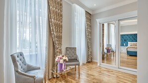 Una habitación amplia con un gran ventanal, un sillón capitonado, otro sillón más pequeño y una mesa de centro de vidrio con un jarrón de flores.