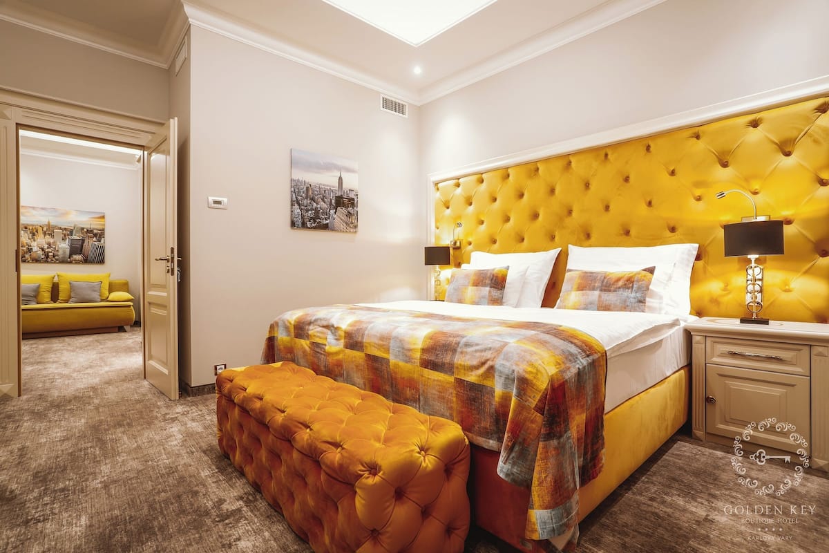 Habitación de hotel con una cama grande, un cabecero amarillo, un banco acolchado y una pintura de un paisaje urbano en la pared.