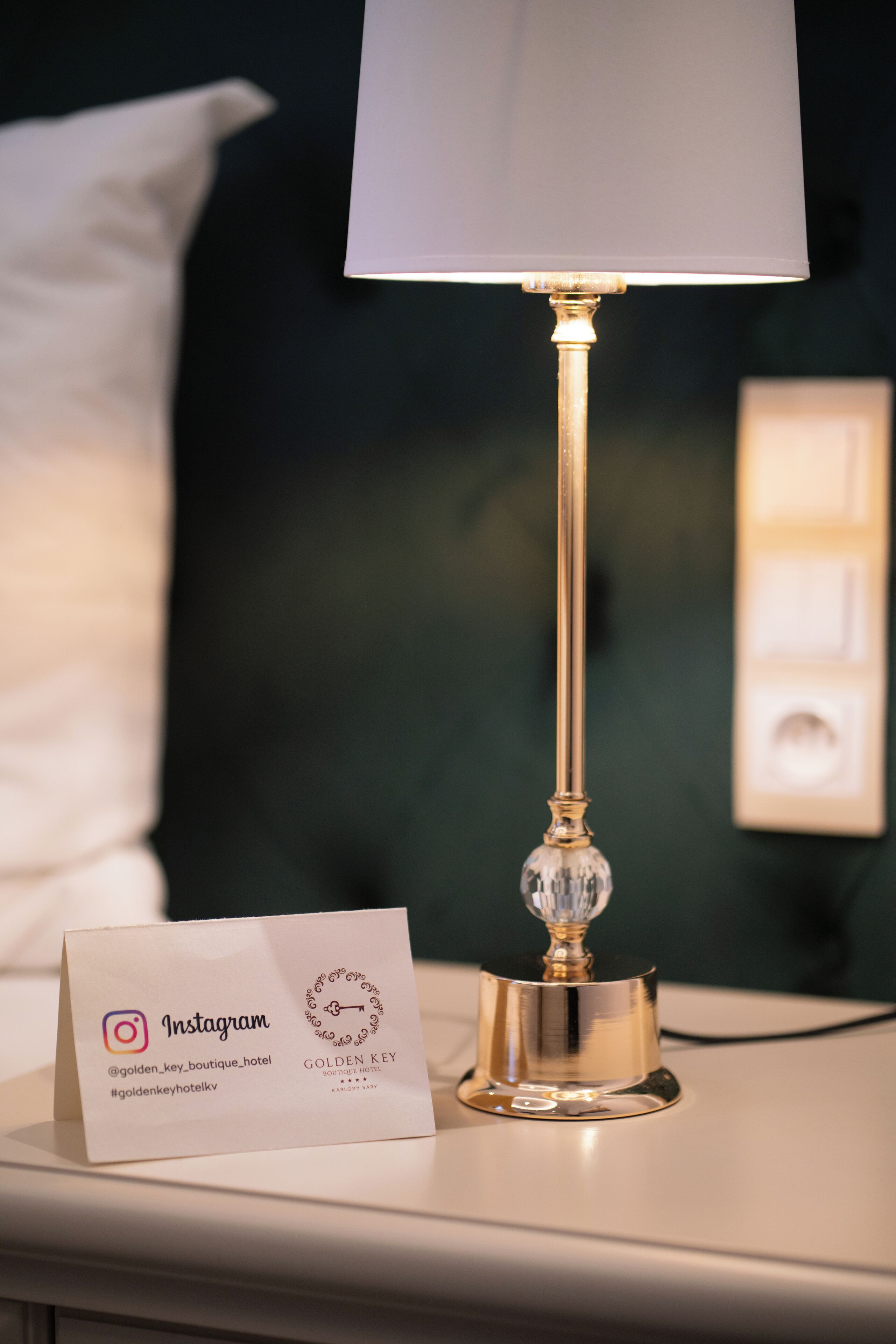 Une table de chevet avec une lampe, un oreiller blanc et une carte de visite.