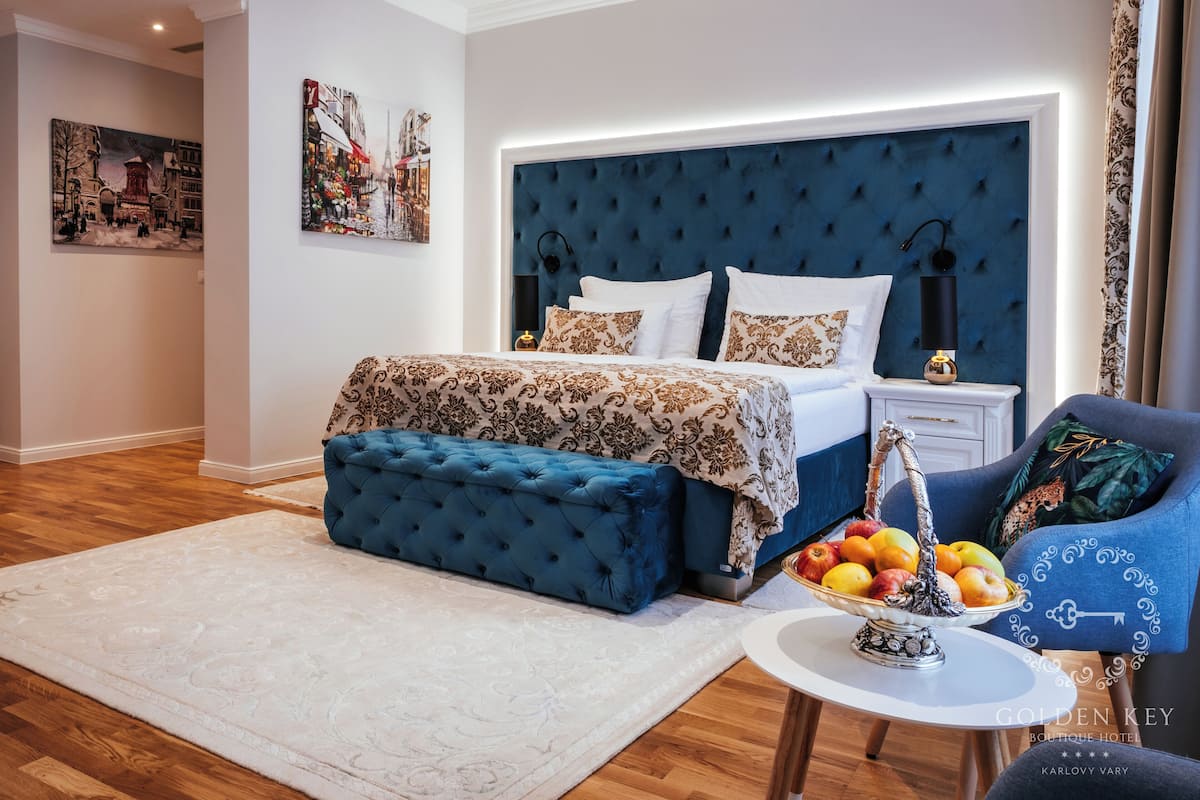 Una habitación de hotel moderna con una cama grande, un banco tapizado en azul, una mesa redonda con fruta y obras de arte en las paredes.