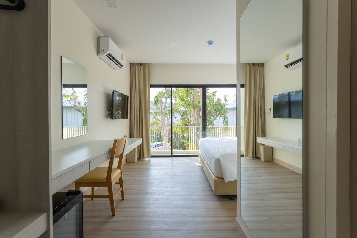 A hotel room with a bed, a desk, a chair, a television, and a balcony with a view of trees and a fence.