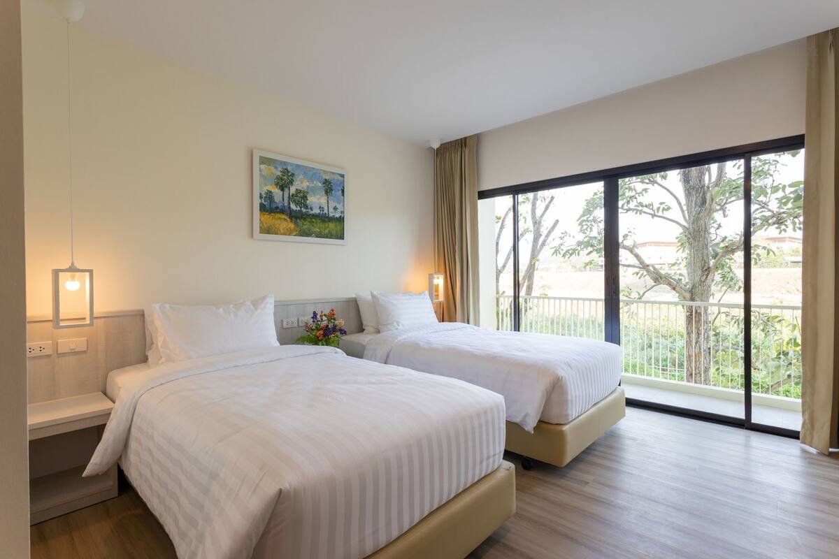 A hotel room with two beds, a painting on the wall, and a balcony with a view.
