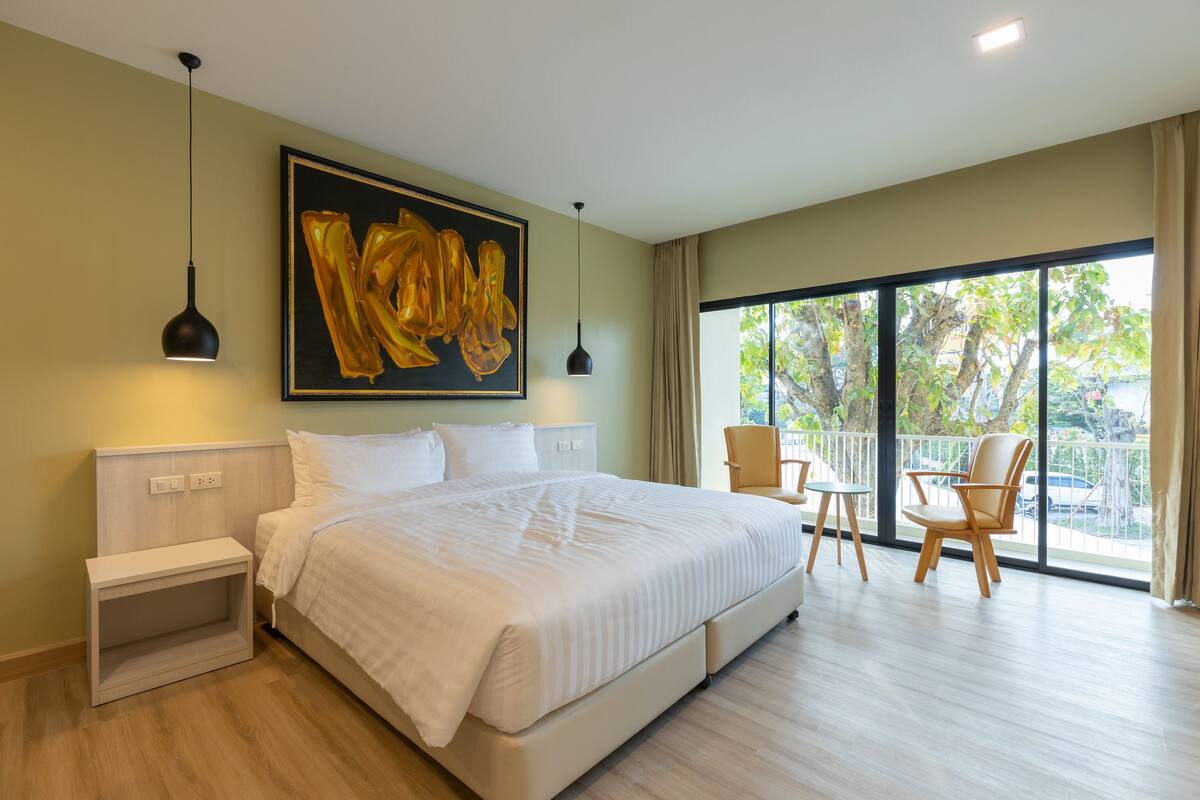 A modern hotel room with a large bed, bedside tables, a painting, and a balcony with chairs.