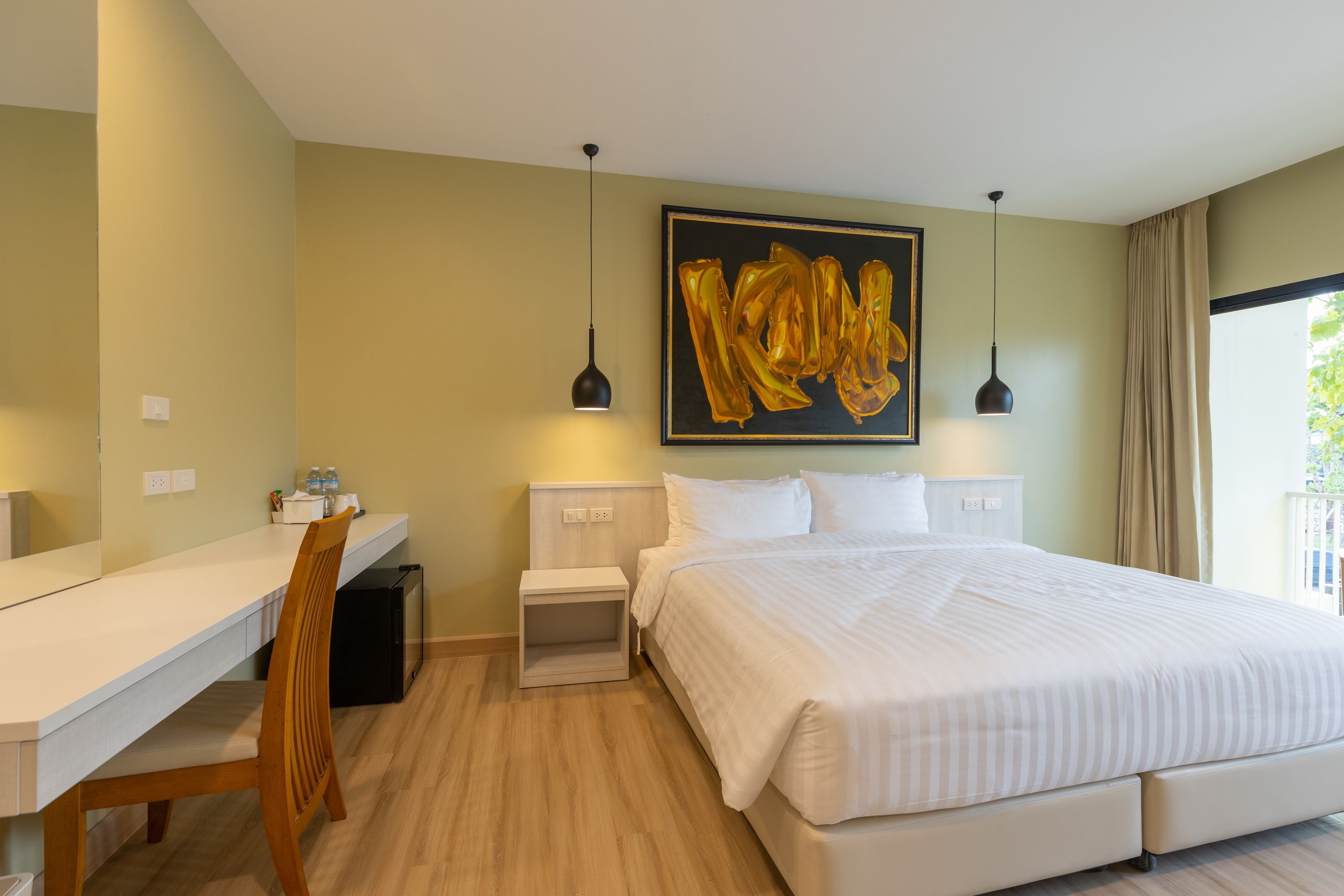 A hotel room with a large bed, a desk, and a chair. There is a painting on the wall and a window with curtains.