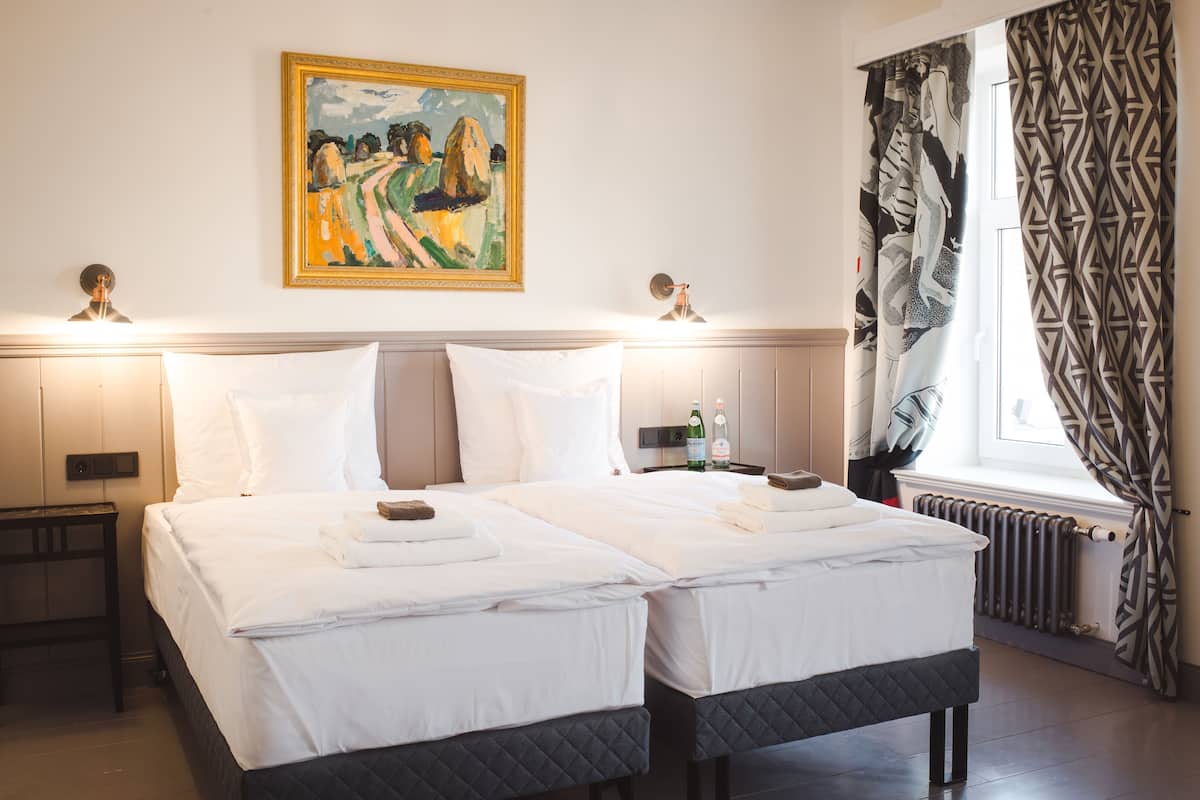Habitación de hotel con dos camas, un cuadro en la pared, una ventana con cortinas estampadas y una mesita de noche con una botella y vasos.