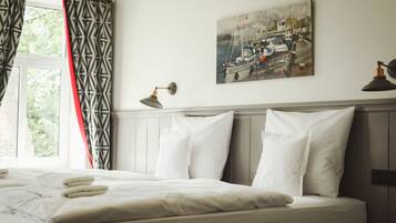 A hotel room with a neatly made bed, a painting of a harbor scene, and a lamp on the nightstand.
