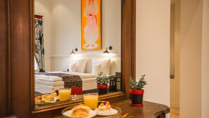 Habitación de hotel con cama, mesita de noche con bandeja de desayuno, espejo que refleja la habitación, un cuadro en la pared y una planta en maceta.