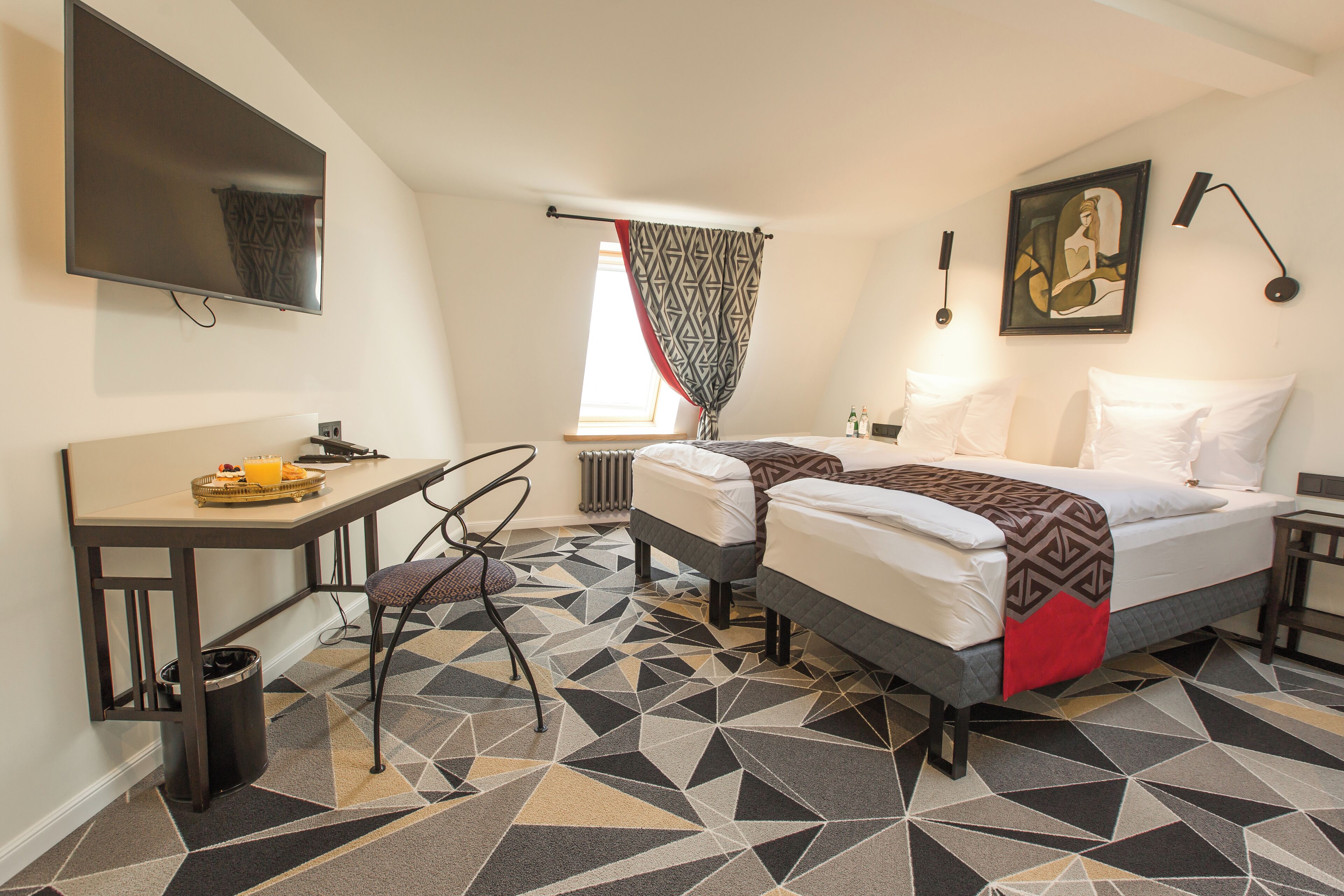 A hotel room with a bed, a desk, a chair, a TV, and a patterned floor.