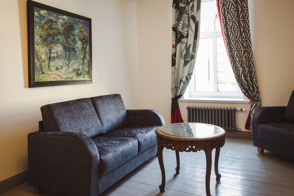 A living room with a dark blue sofa, a glass-top coffee table, and a painting on the wall. - Art Hotel Roma (Liepaja)