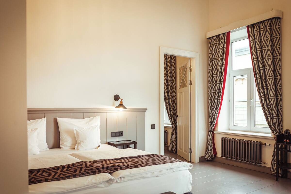 Una cama bien hecha con sábanas blancas, una mesita de noche con una lámpara y una ventana con cortinas estampadas.