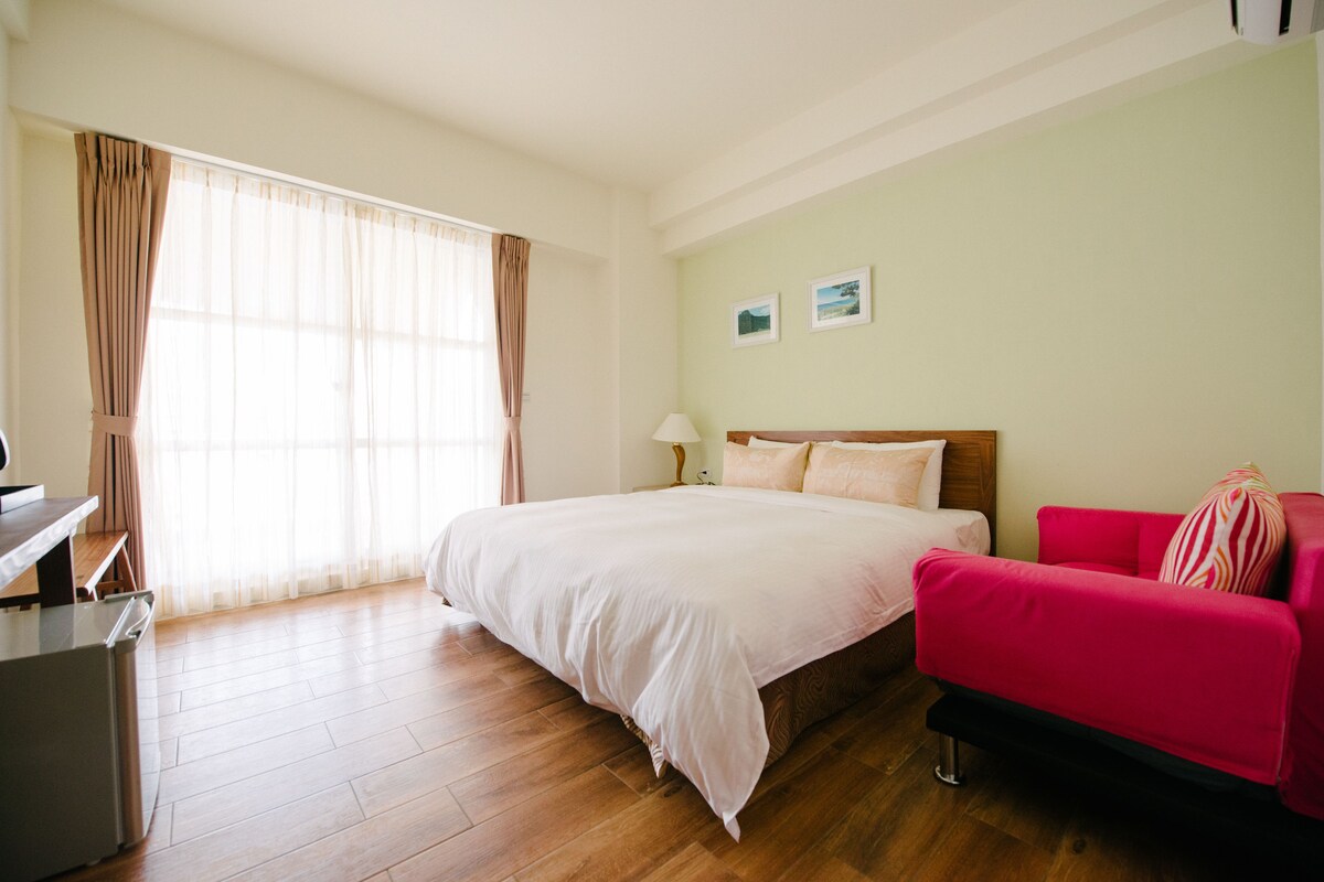 A hotel room with a large bed, a desk, a chair, a red sofa, and a window with curtains.