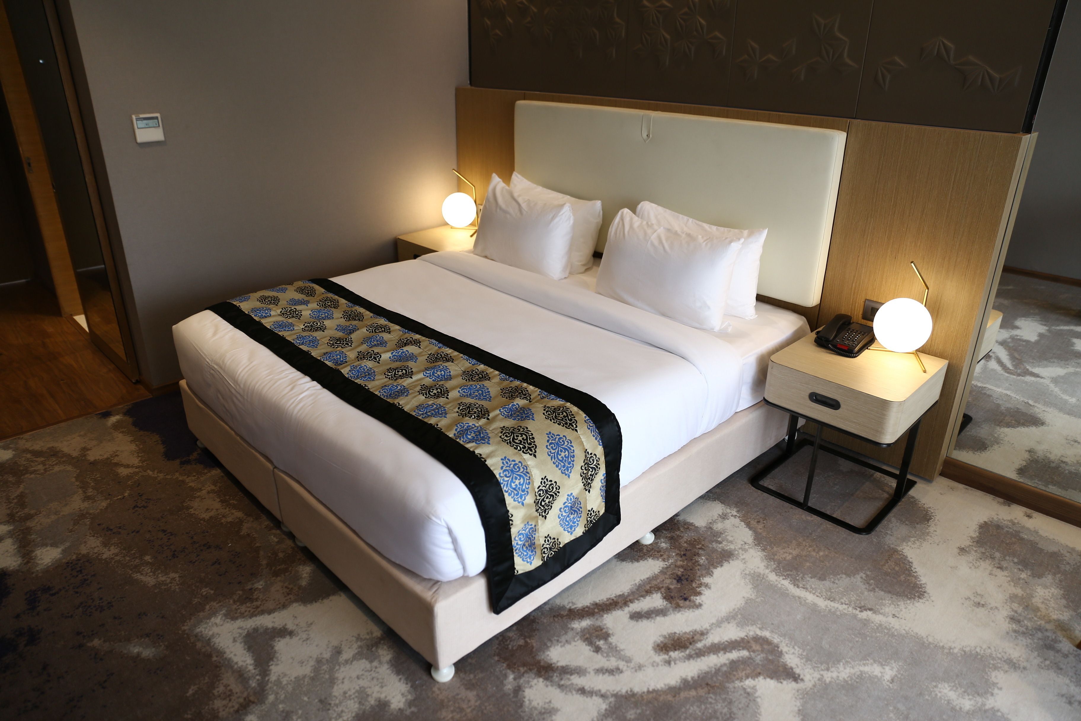 A neatly made bed with a patterned bedspread, two bedside lamps, and a bedside phone on a wooden nightstand.