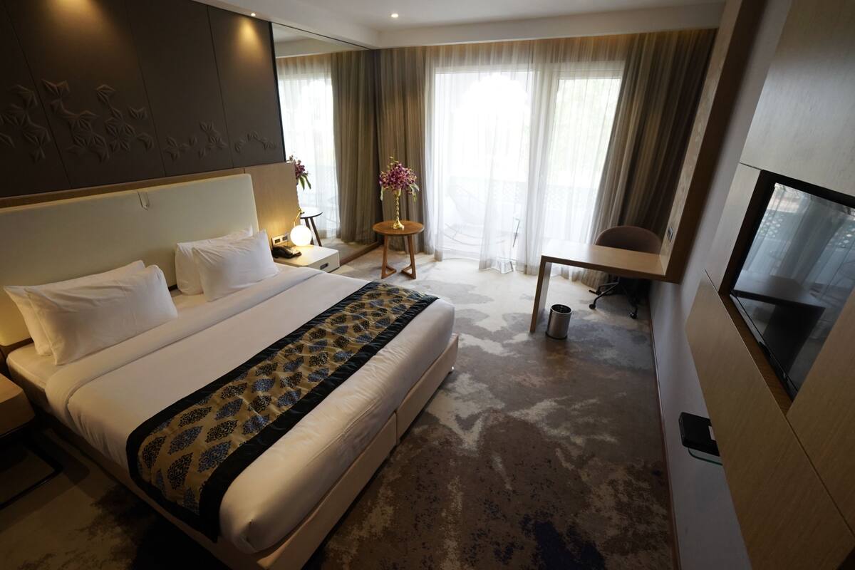 Una habitación de hotel con una cama grande, un escritorio, una silla, un televisor y una pequeña mesa con un jarrón de flores.