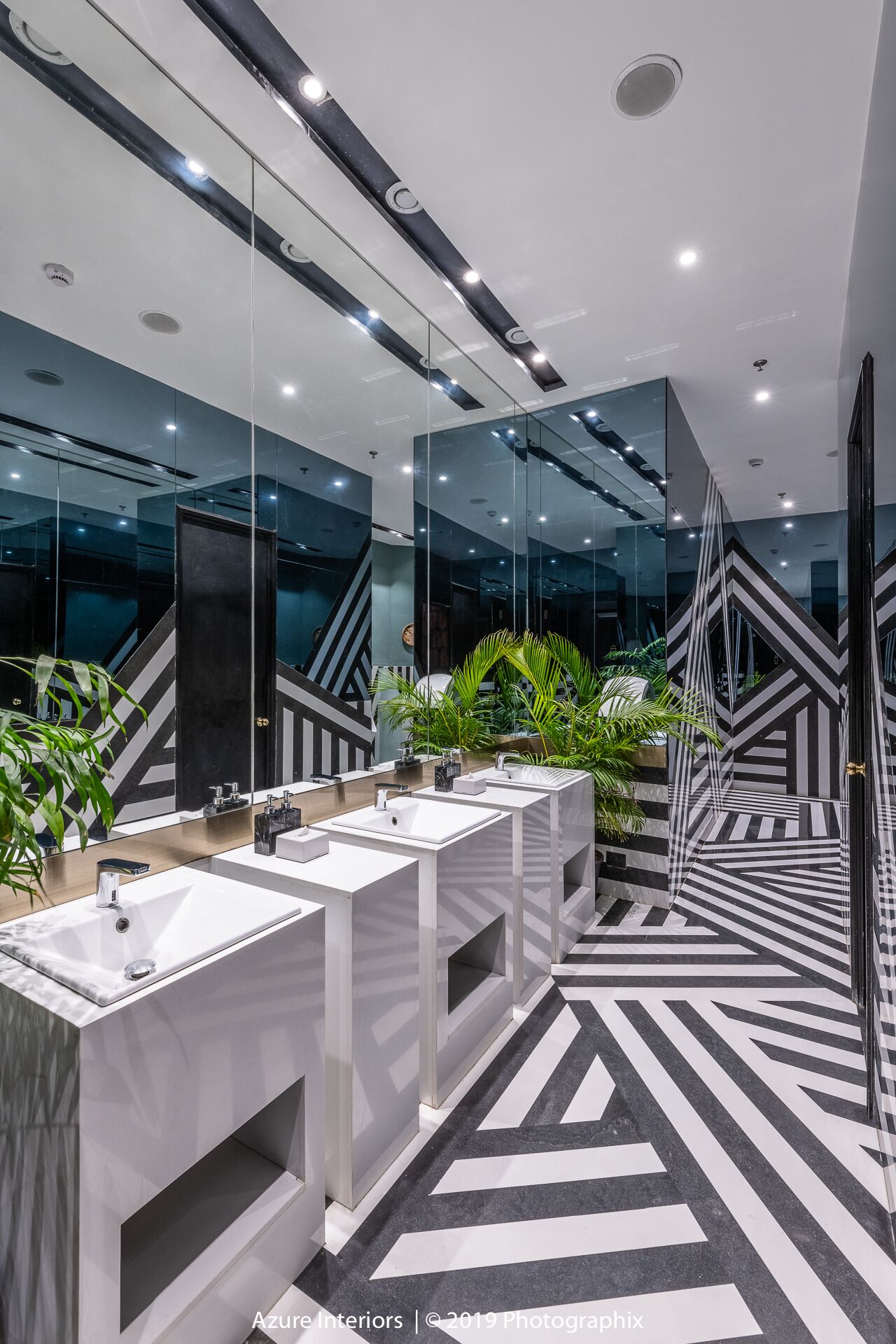 Un baño moderno con paredes espejadas, varios lavamanos y un suelo con estampado geométrico.