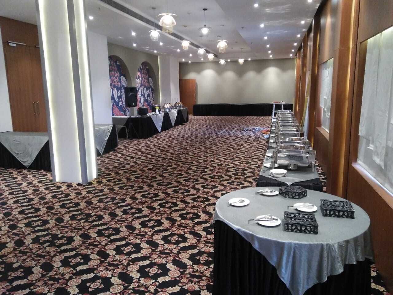 Un salón de conferencias con alfombra estampada, mesas redondas con sillas y un escenario con un orador y una pantalla.