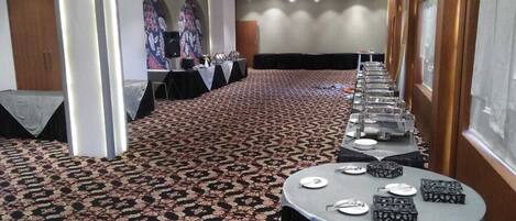Un salón de conferencias con alfombra estampada, mesas redondas con sillas y un escenario con un orador y una pantalla.