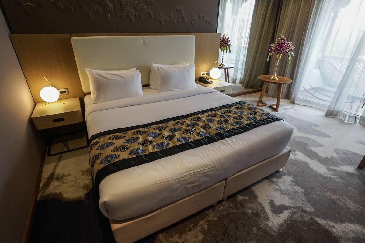 Habitación de hotel con una cama grande, una mesita de noche con una lámpara, una pequeña mesa con un florero y vistas a través de cortinas translúcidas.