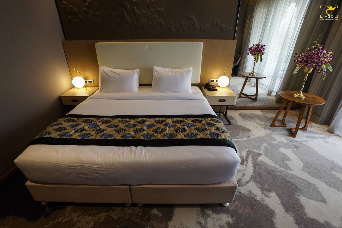 Una habitación de hotel con una cama grande, dos mesitas de noche con lámparas, una mesita auxiliar con flores y un cabecero decorativo.