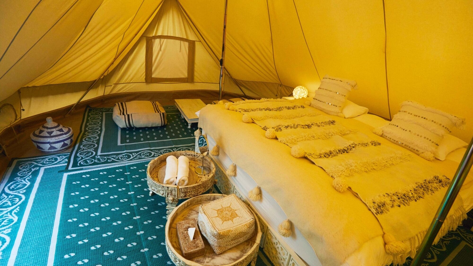 A tent interior with a bed, pillows, a lantern, and a patterned mat.