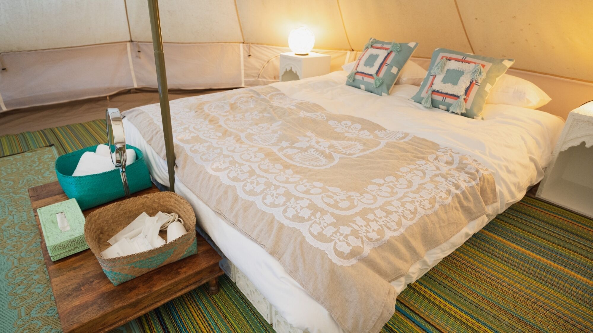 A room with a bed, a bedside lamp, a nightstand, a basket, and a small table with a tissue box.