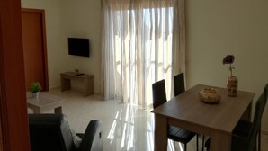 A spacious living room with a wooden dining table, black chairs, a TV on a stand, and a large window with sheer curtains.