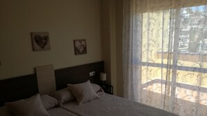 A hotel room with two beds, a headboard, a nightstand with a lamp, and a window with sheer curtains.
